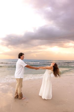 Romantic couple holding hands and spinning on sandy beach at sunset with dramatic cloudy sky backdrop.
