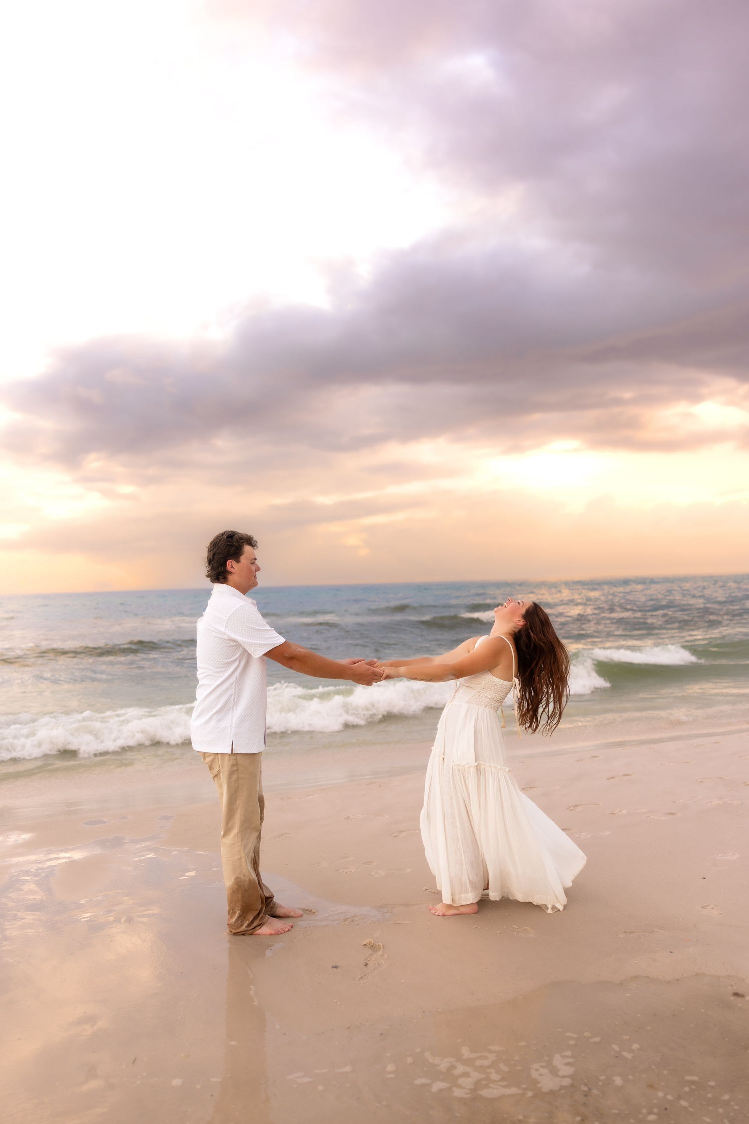 Couple holding hands and dancing playfully on a sandy beach at sunset with dramatic cloudy sky.
