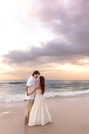 Romantic couple holding hands and nearly kissing on beach shoreline beneath a dramatic sunset sky.