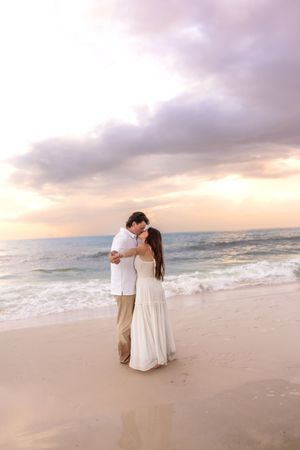 Couple kissing on sandy beach at sunset with ocean waves behind them.