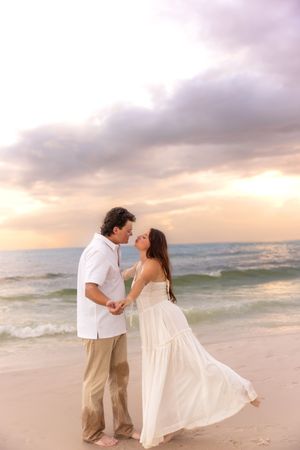 Romantic couple holding hands and almost kissing on a beach at sunset, woman in flowing white dress.