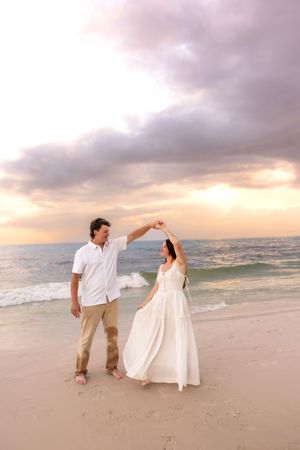 Man spinning woman in white dress on sandy beach during a beautiful colorful sunset.