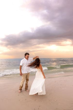Couple dancing on beach at sunset, woman in white dress spinning as waves crash behind them.