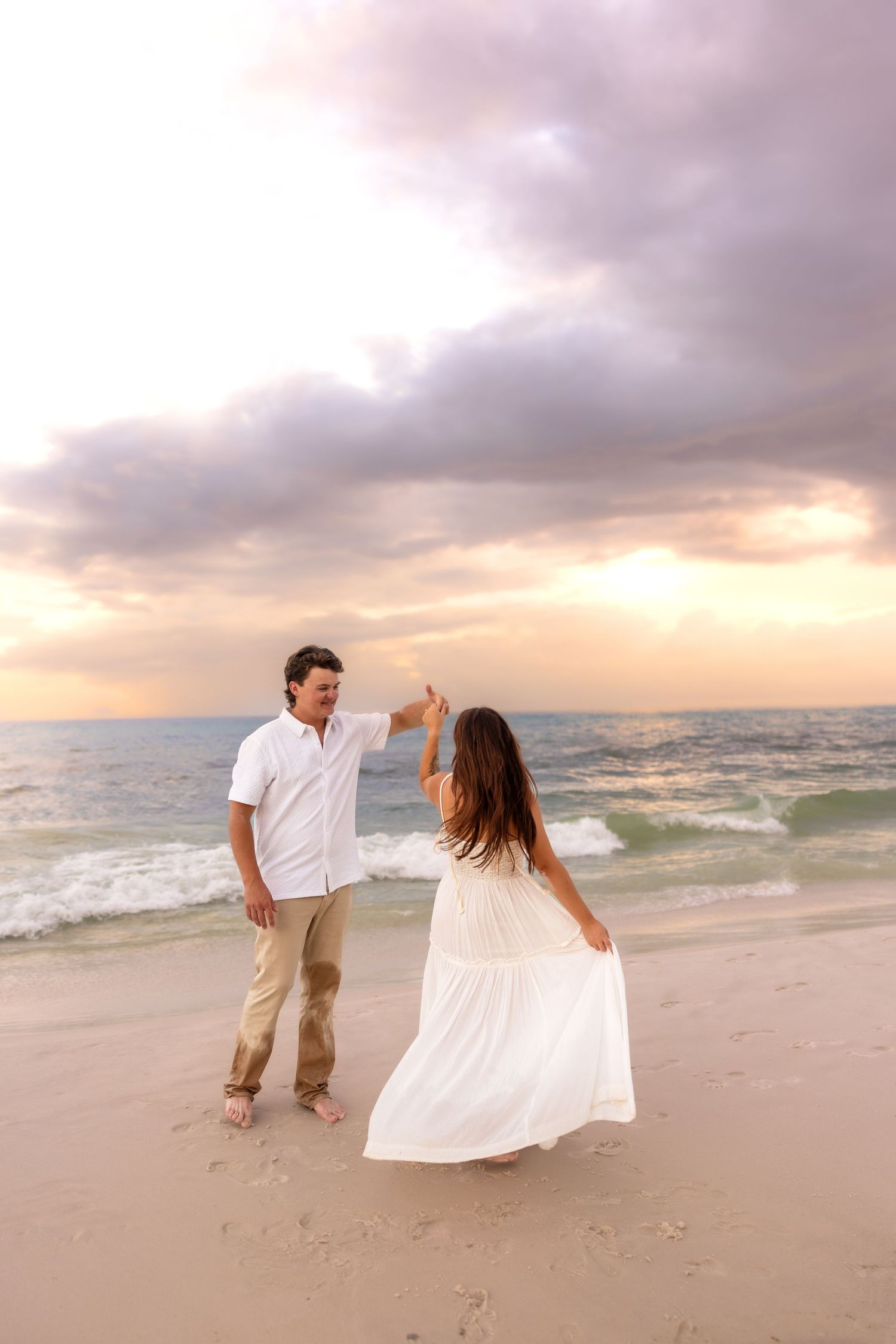 Man spinning woman in white dress on a beach at sunset, waves and colorful sky in background.
