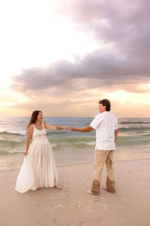 Couple holding hands on a sandy beach at sunset, wearing white outfits with ocean waves in the background.