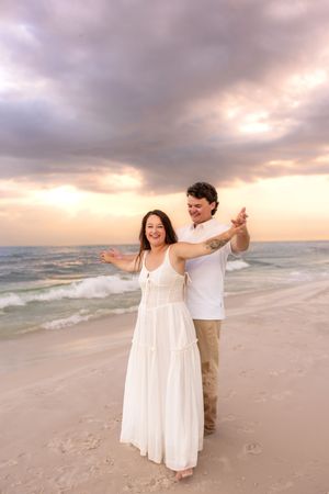 Joyful couple dancing with arms outstretched on beach at sunset, woman wearing flowing white dress.