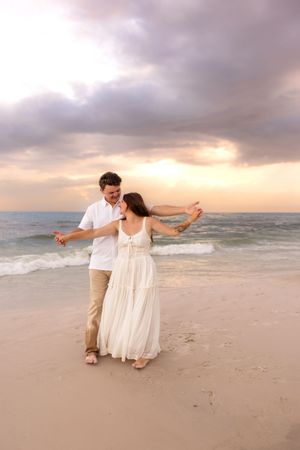 Happy couple dancing together on sandy beach at sunset, woman in white dress twirling joyfully.