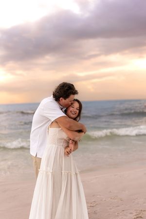Man hugging woman in white dress from behind on a beach at sunset, both smiling happily.