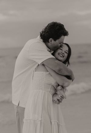 Black and white photo of man embracing woman in white dress on beach, both smiling joyfully.