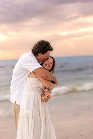 Couple embracing on beach at sunset, woman in white dress laughing joyfully as man hugs her from behind near ocean waves.