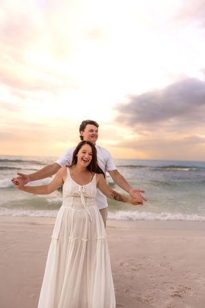 Joyful couple posing on beach at sunset, woman spreading arms wide.