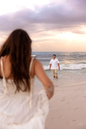 Woman in white dress reaching toward man walking out of ocean waves at a colorful beach sunset.