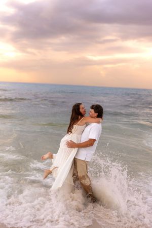 Man lifting woman in white dress while ocean waves splash around them at sunset.