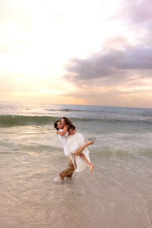 Man carrying woman in white dress through ocean waves at sunset on a beautiful beach.