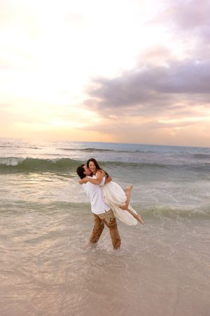 Man lifting woman in white dress while standing in ocean waves on a beach at sunset.