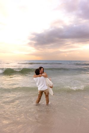 Man carrying woman in his arms while wading in ocean waves at a colorful beach sunset.