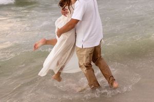 Couple walking barefoot through ocean waves, woman in flowing white dress held by man in khakis.