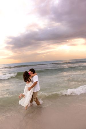 Romantic couple kissing while wading in ocean waves at sunset, woman wearing flowing white dress on sandy beach.