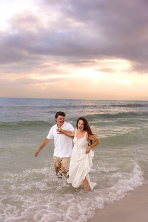 Joyful couple laughing and running through ocean waves on a sandy beach at sunset.