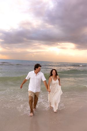 Happy couple in white holding hands walking through ocean waves on a sandy beach at sunset.