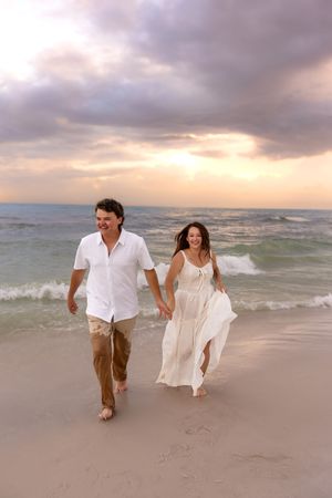 Couple holding hands running barefoot on sandy beach at sunset, woman in white dress, man in white shirt.
