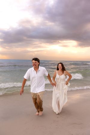 Couple in white outfits walking hand in hand on a sandy beach at sunset with ocean waves behind them.