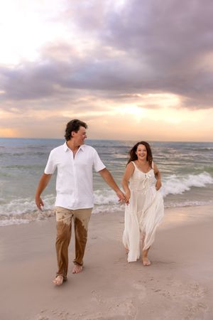 Couple holding hands walking on a sandy beach at sunset, woman in white dress, man in white shirt.