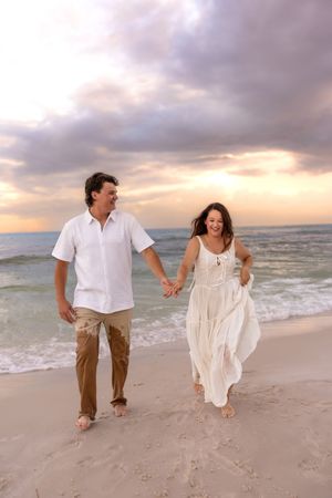 Couple holding hands walking barefoot on sandy beach at sunset, woman in white dress, man in white shirt and khaki pants.