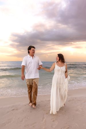 Couple holding hands walking on sandy beach at sunset, woman in white dress and man in white shirt.