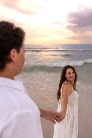 Couple holding hands on beach at sunset, woman in white dress smiling back.