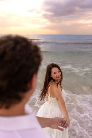 Smiling woman in white dress holds hands with partner on beach at sunset.