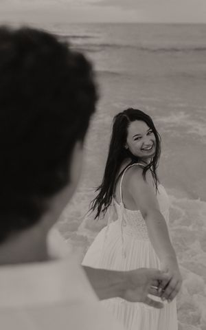 Black and white photo of a smiling woman in a white dress holding hands with a man on a beach.