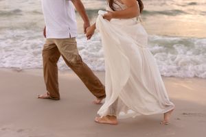 Barefoot couple walking on a sandy beach, woman in flowing white dress and man in khaki pants, waves in background.