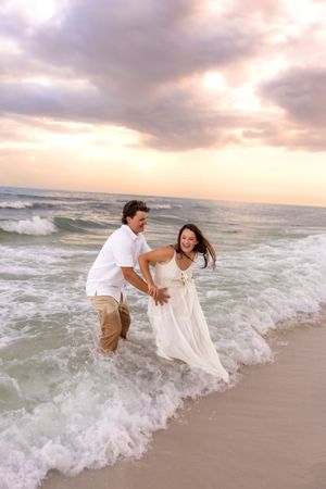 Couple laughing and running through ocean waves at sunset on a beach, woman in white dress.