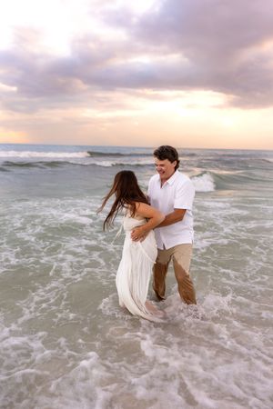 Couple dancing in ocean waves at sunset, woman in white dress, man in white shirt, romantic beach photography.