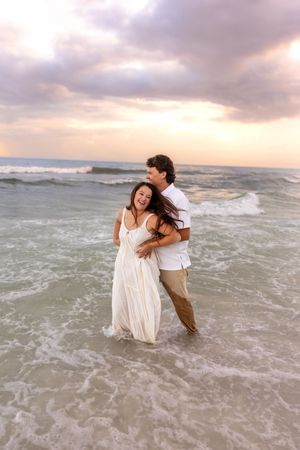 Couple laughing and embracing in ocean waves at sunset, woman in white dress, man in white shirt and khakis.