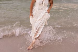 Woman in white boho maxi dress walking barefoot on sandy beach with ocean waves.