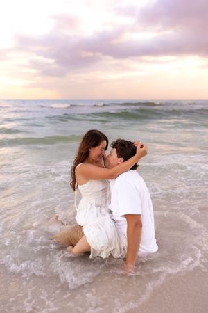 Romantic couple embracing in ocean waves on the beach at sunset, both wearing white clothing.