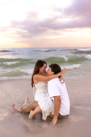 Romantic couple sitting in ocean waves on a beach at sunset, embracing and almost kissing.