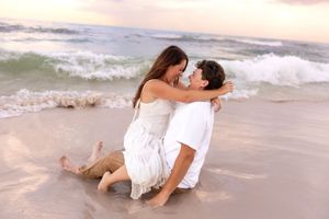Romantic couple embracing on a sandy beach with ocean waves, sitting close together in white outfits at sunset.