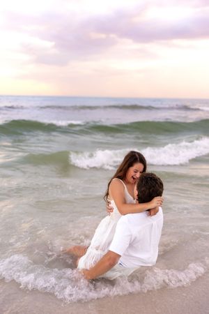 Couple embracing joyfully in ocean waves at sunset, both wearing white outfits on a tropical beach.