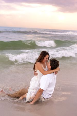 Romantic couple embracing in ocean waves at sunset, woman in white dress sitting on man's lap on wet sandy beach.