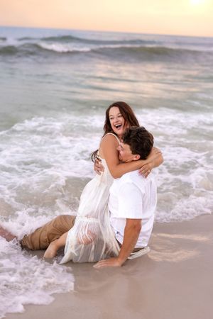 Couple laughing and embracing in ocean waves on a beach at sunset, wearing white outfits.
