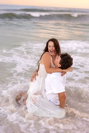 Joyful couple embracing in ocean waves at sunset, woman in white dress laughing as man kneels in the surf.