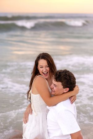 Joyful couple laughing and hugging in ocean surf at sunset, woman in white dress and man in white shirt.