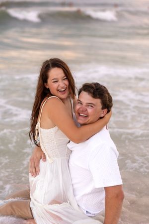 Happy couple sitting in ocean waves laughing, woman in white dress hugging man in white shirt at beach.