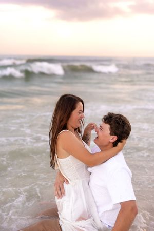 Happy couple sharing a tender moment in shallow ocean waves at sunset, woman in white dress embracing man.