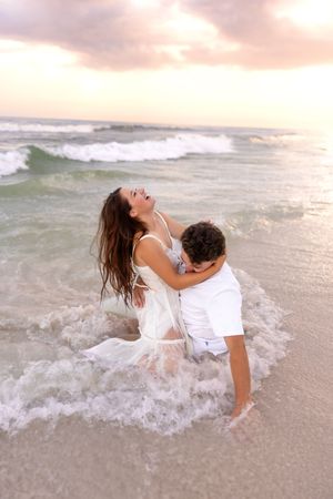 Man lifting woman in white dress in ocean waves at sunset, both laughing joyfully on beach.