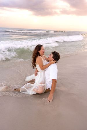Romantic couple sitting together in beach waves at sunset, both dressed in white, smiling at each other.