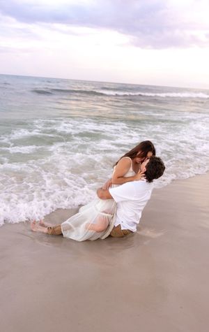 Romantic couple embracing and kissing in ocean waves on beach, both wearing white outfits.